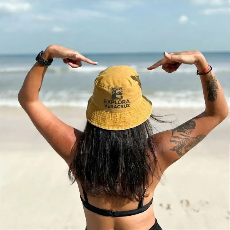 Vista posterior de una mujer en la playa señalando un sombrero tipo bucket color mostaza con el logotipo de 'Explora Veracruz', con el mar y la arena de fondo.