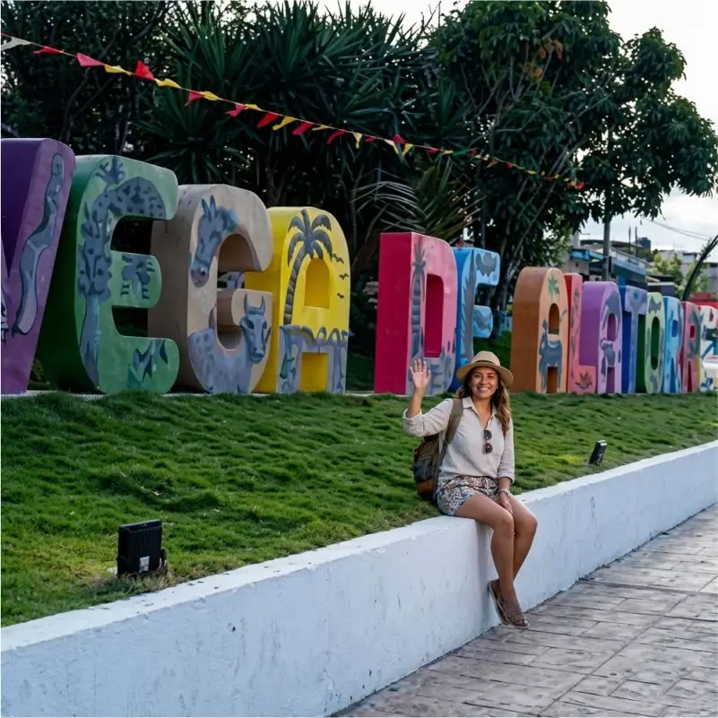 Turista sonriendo y saludando sentada junto a las letras monumentales de colores de 'Vega de Alatorre', Veracruz. La turista lleva un sombrero de paja, mochila y blusa beige. Las letras están decoradas con murales de flora y fauna local y una palmera, sobre césped y rodeadas de vegetación y banderas