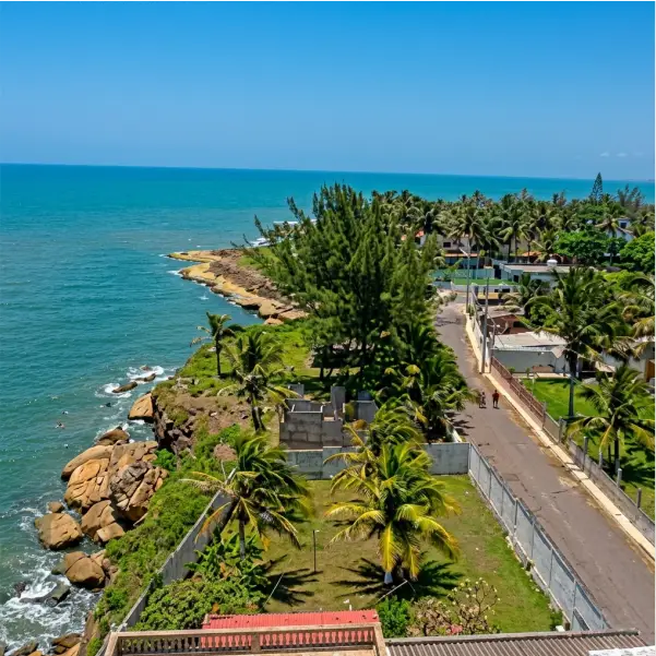 Un primerísimo plano de una costa rocosa y una calle residencial elevada en Cazones de Herrera, Veracruz. La imagen muestra la orilla del mar turquesa a la izquierda, una pared de concreto a la derecha que separa propiedades llenas de palmeras y vegetación tropical, y una calle pavimentada donde caminan dos figuras. Al fondo, se aprecia un tramo de arena y más vegetación.
