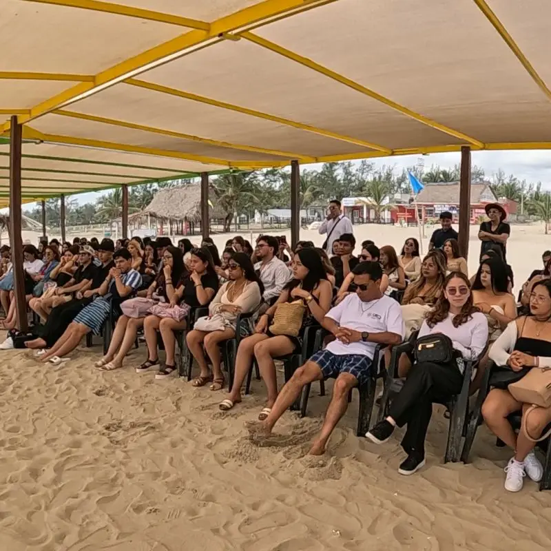 Un grupo numeroso de jóvenes estudiantes y turistas sentados en filas de sillas sobre la arena bajo una gran estructura de sombra en la playa, asistiendo a una plática de educación ambiental en el Campamento Tortuguero Mar de Plata en Tuxpan, Veracruz.