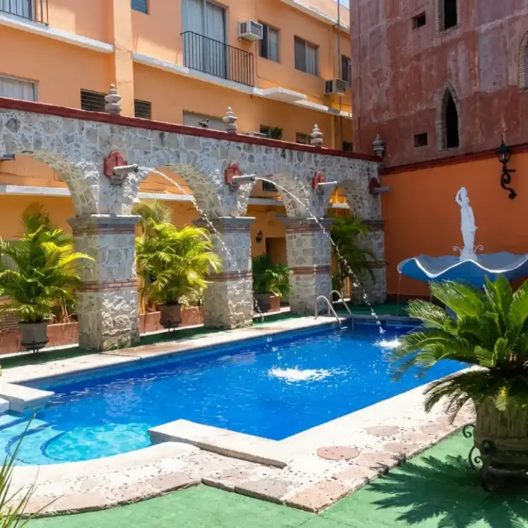 Piscina del Hotel Tajín Papantla con fuentes de agua cayendo desde arcos de piedra, vegetación tropical y estatua decorativa.