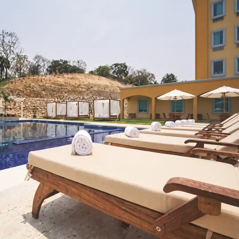 Alberca al aire libre con asoleadero, camastros de madera y exclusivas cabañas en el Hotel La Quinta by Wyndham Poza Rica, un oasis urbano para el descanso del viajero.