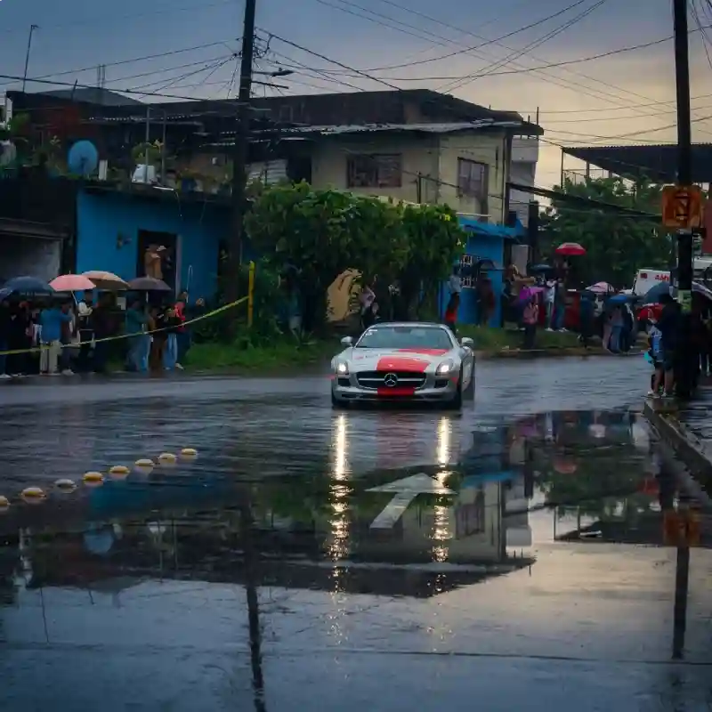 Auto deportivo circulando bajo la lluvia por una calle poblada con espectadores protegidos con paraguas en Tlapacoyan durante La Carrera Panamericana.