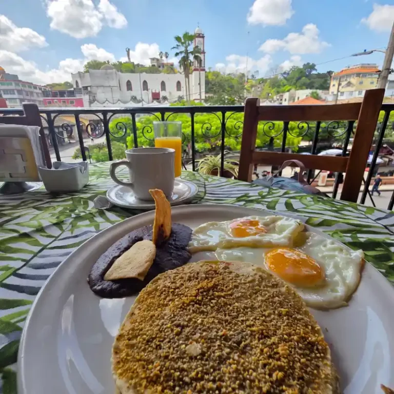 Desayuno completo con empipianadas tradicionales, huevos estrellados, frijoles negros con totopo, taza de café con vainilla y jugo de naranja en la terraza del Restaurante Plaza Pardo con vista a la parroquia de Papantla.