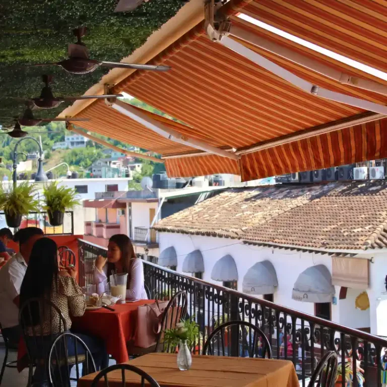 Turistas conversando en la terraza elevada del Restaurante Plaza Pardo en Papantla bajo un amplio toldo a rayas, rodeados de techos de teja colonial y vegetación en el balcón.