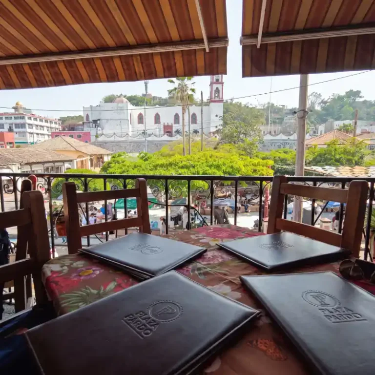 Vista aérea en contrapicado de drone sobre el Zócalo de Papantla, Veracruz, capturando el edificio colonial histórico que alberga el Restaurante Plaza Pardo y su terraza con toldos, rodeado de calles adoquinadas y las colinas de la costa.